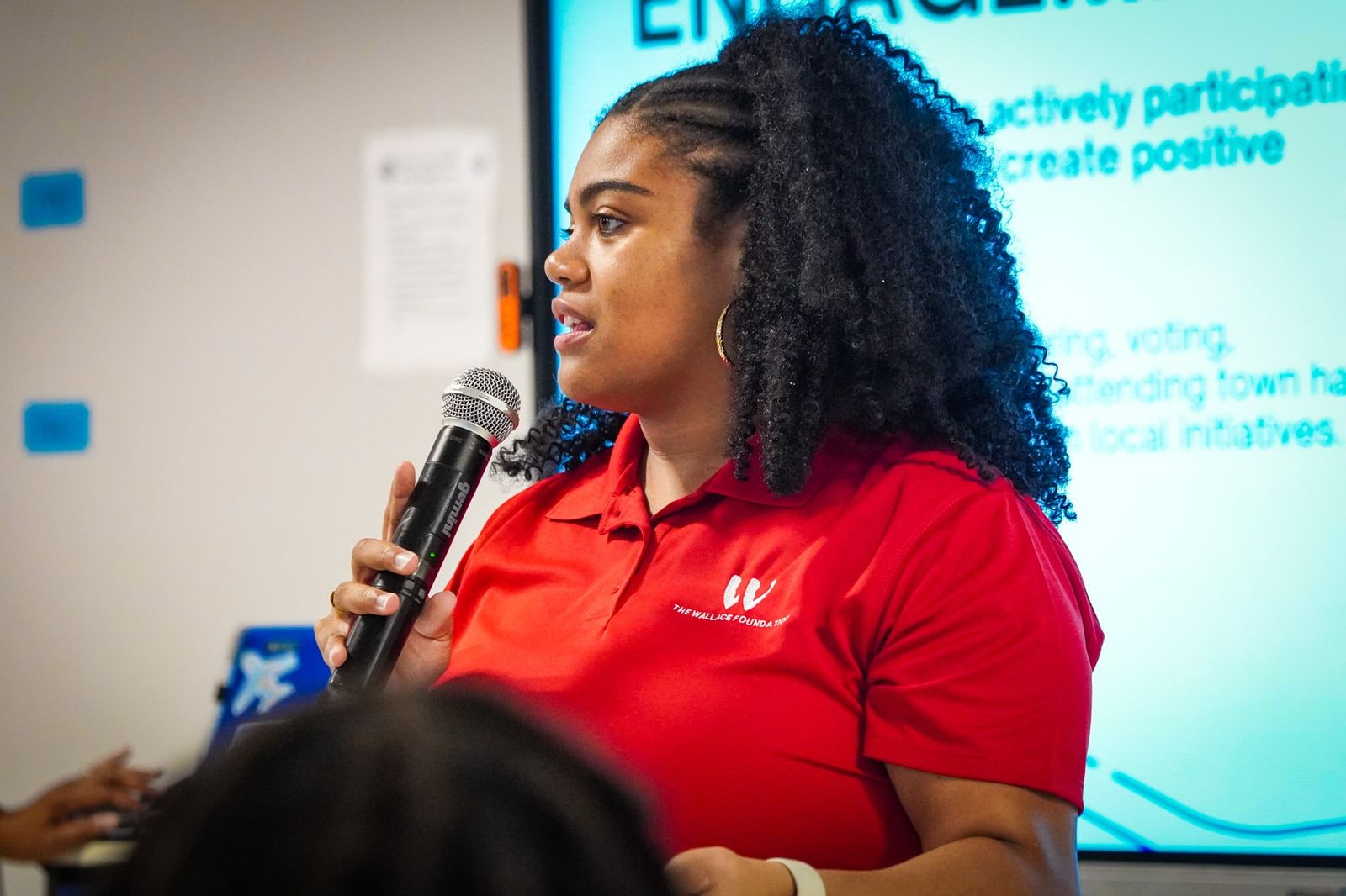 A Foundation team member in a red Wallace Foundation polo speaking into a microphone beside a projected slide on civic engagement, listing voting, town halls and local initiatives.