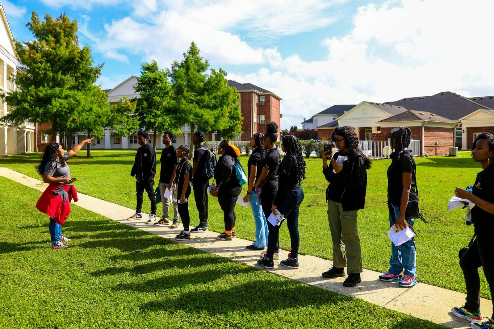 A guide pointing out a building while a line of students listens on a campus walkway, several in Wallace Foundation shirts and holding tour papers, with residence halls and trees behind them.