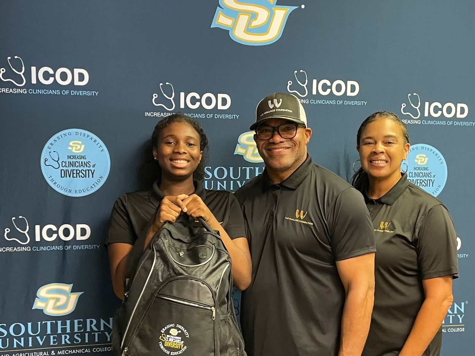 A student holding a drawstring bag, standing between two adults in Wallace Foundation polo shirts in front of a sponsor backdrop.