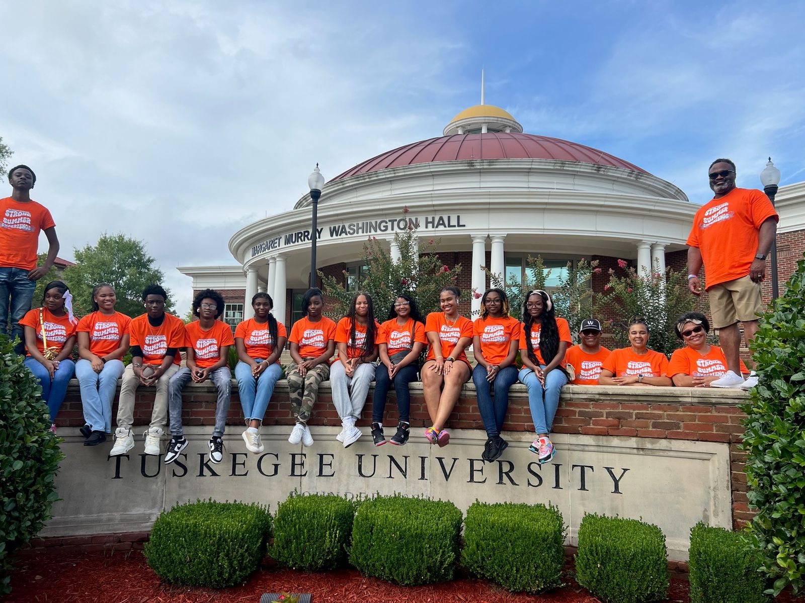 Strong Summer Experience participants and staff in program T-shirts, seated along a low wall beside the Tuskegee University campus sign.