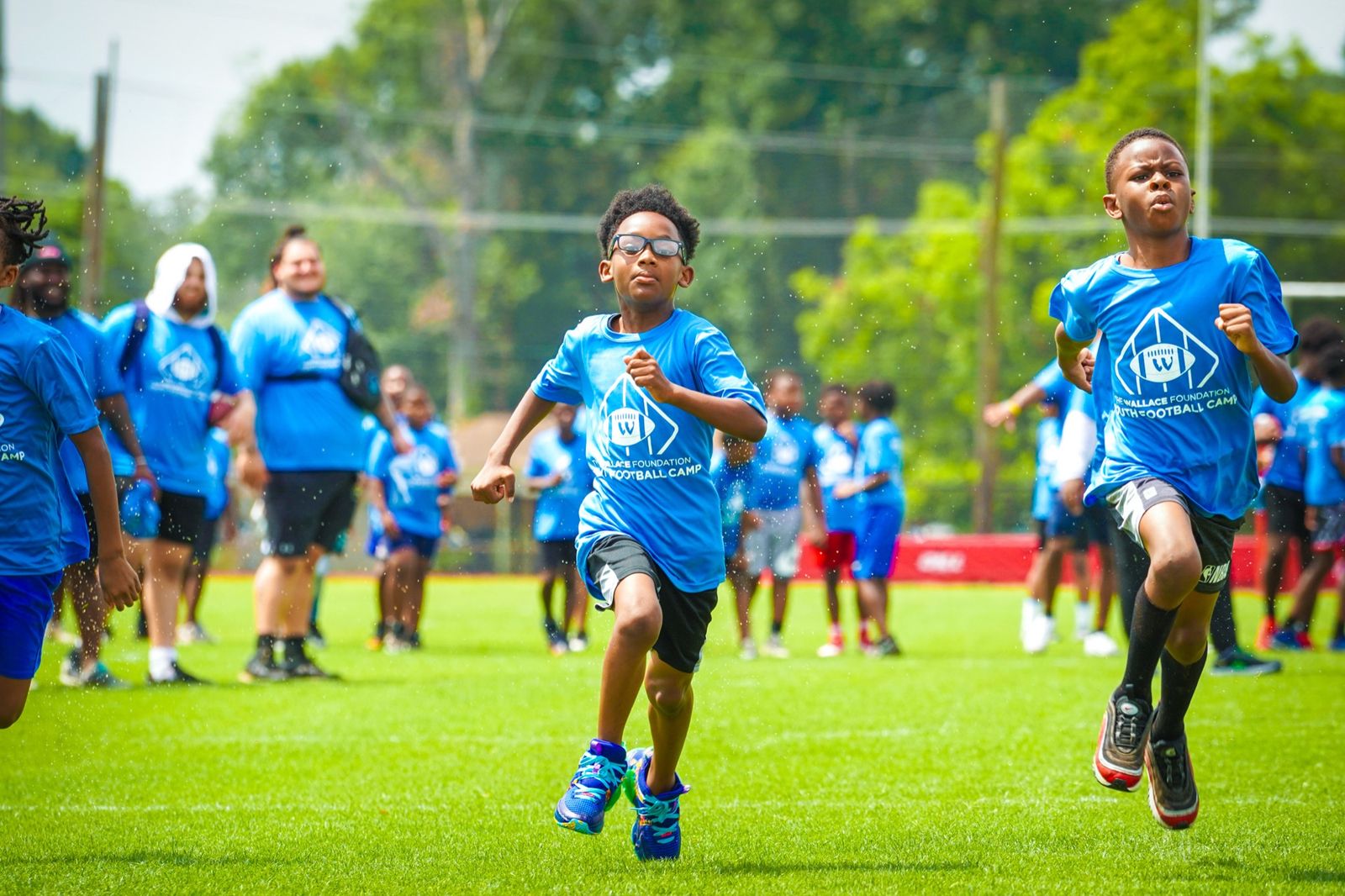 Two boys in bright blue Wallace Foundation Youth Football Camp T-shirts sprinting across a grass field through a spray of water, with other campers and staff in matching shirts behind them.